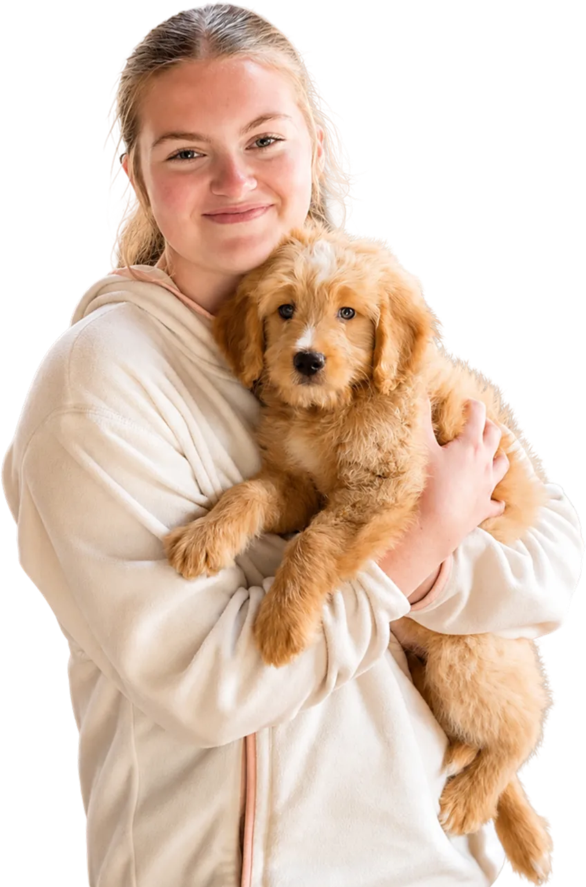 A woman wearing a light-colored fleece jacket holds a fluffy, light brown puppy while smiling at the camera.