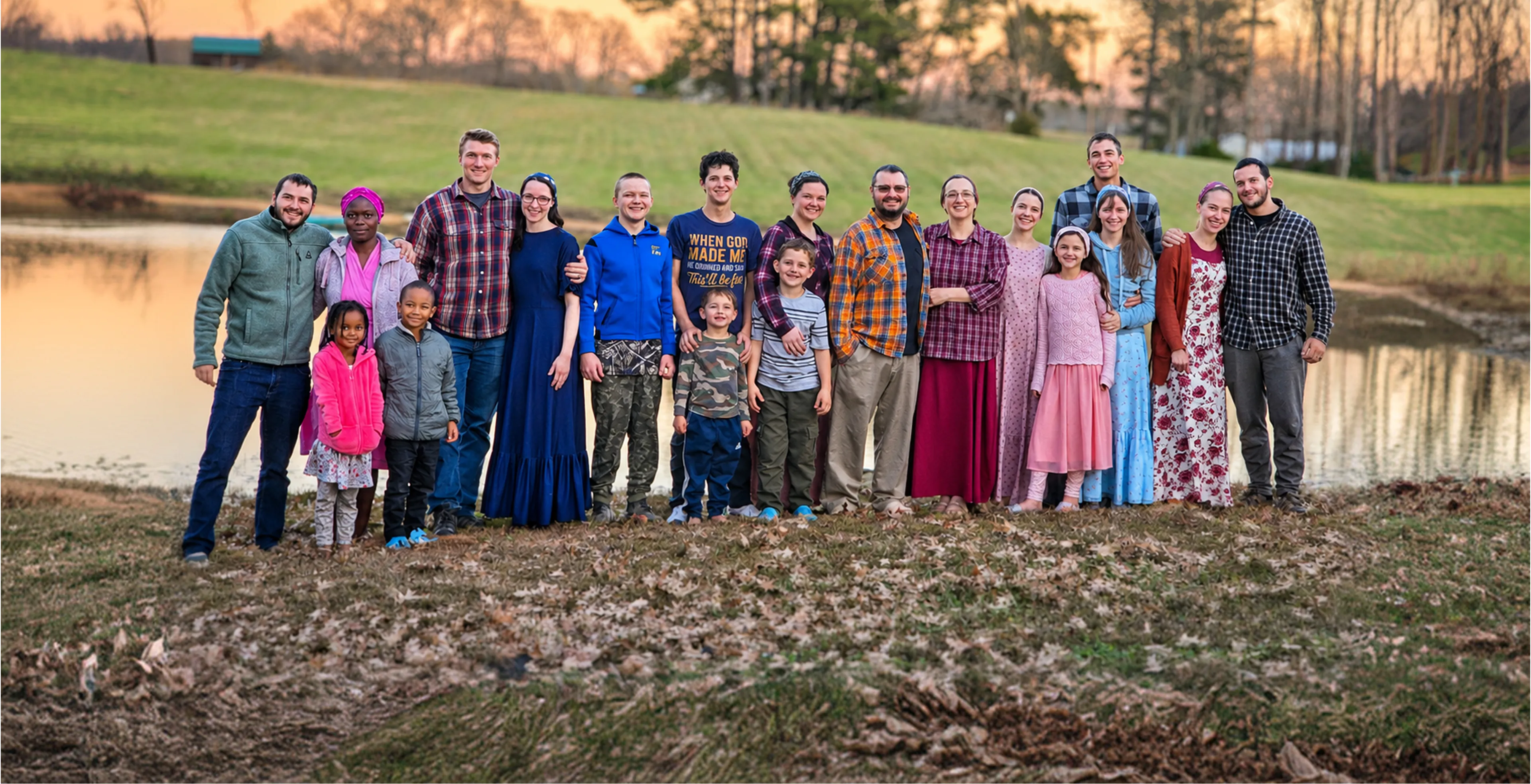 The Carrier Family, including adults and children, stand together outdoors near a pond at sunset, posing for a group photo.