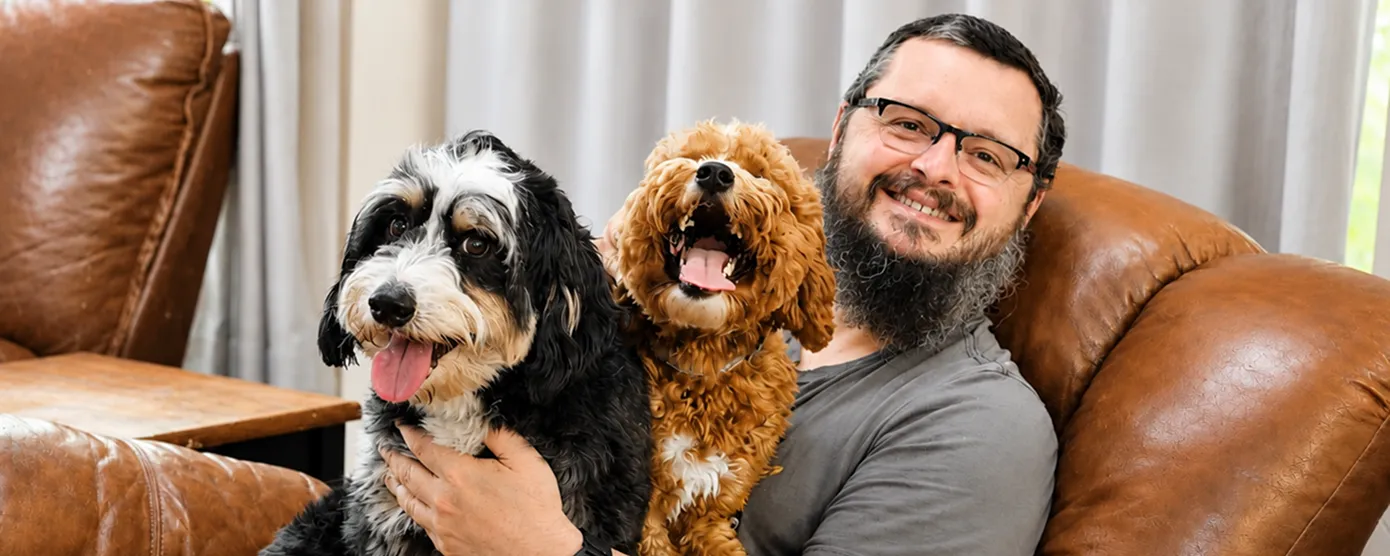 Marc sitting on a brown leather chair smiles while holding two happy dogs, one black and white and one brown, on his lap.