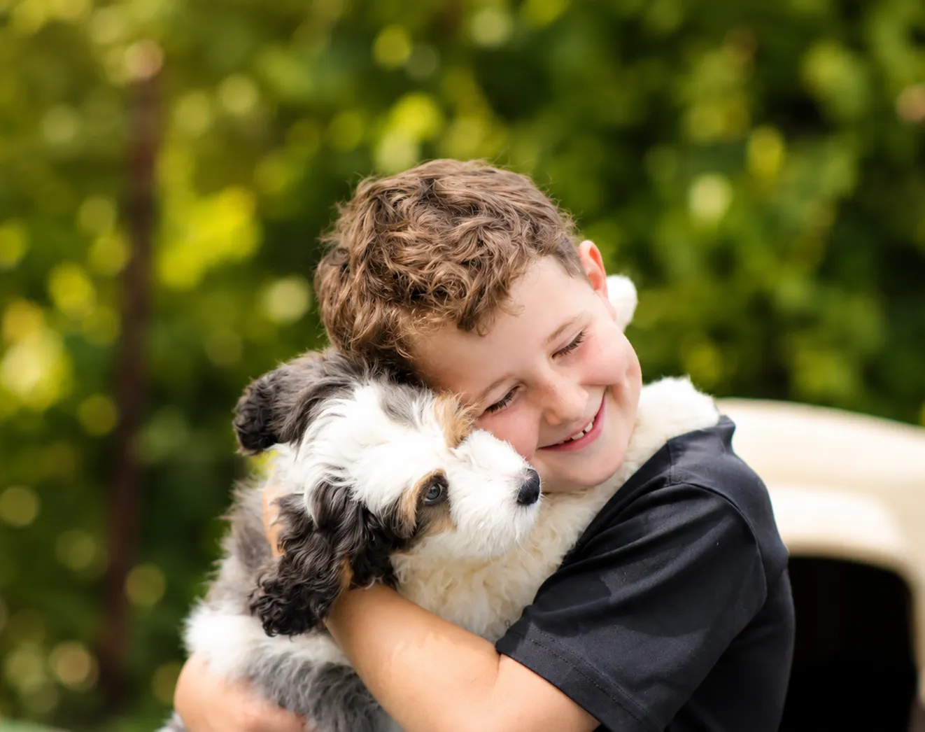 Smiling young boy hugs a fluffy, black and white puppy outdoors.