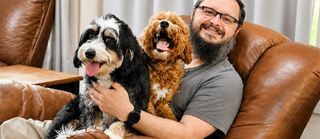 Marc sitting on a brown leather chair smiles while holding two happy dogs, one black and white and one brown, on his lap.
