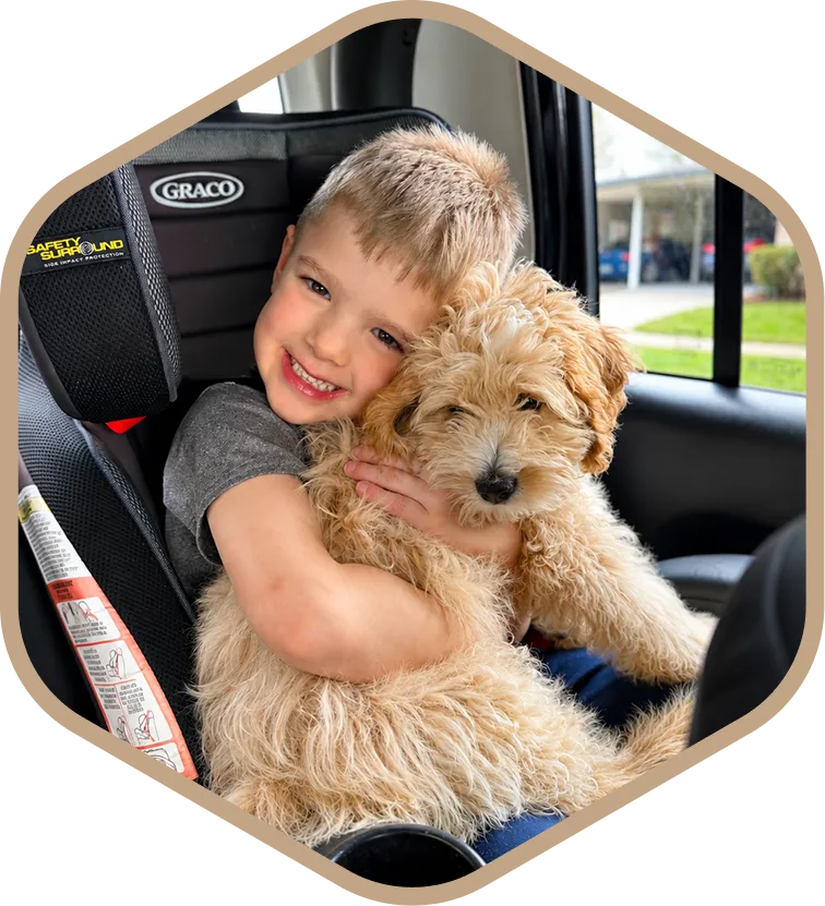 Young boy sitting in a car seat, smiling and hugging a fluffy, light-colored dog.