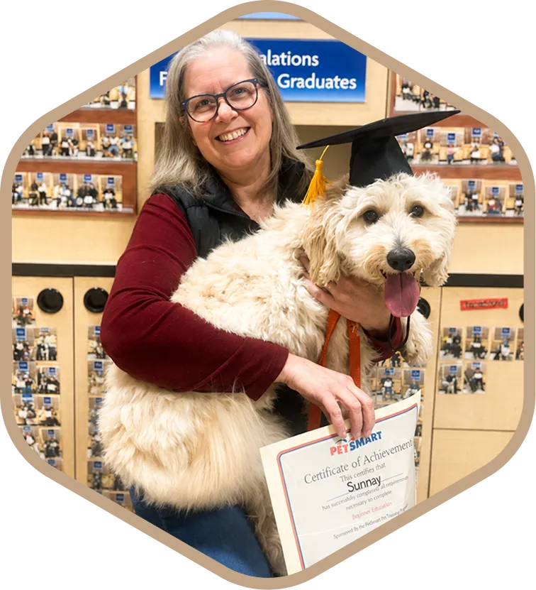 A woman holds a fluffy dog wearing a graduation cap and holding a PetSmart certificate of achievement.