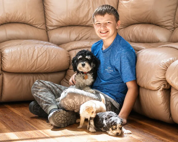 enoch sits on a wooden floor in front of a tan sofa, smiling, with a small black and white dog in his lap and three puppies on the floor beside him.