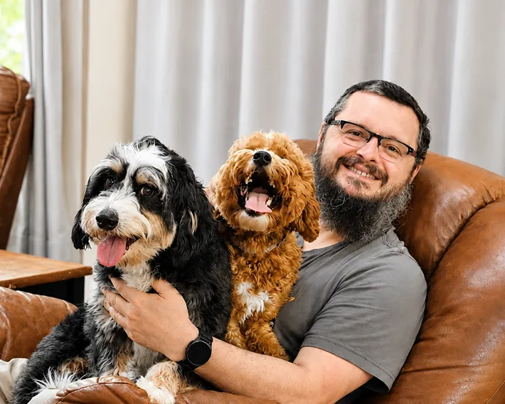 Marc sits on a brown leather chair, smiling and holding two happy dogs, one black and white, the other brown and curly.