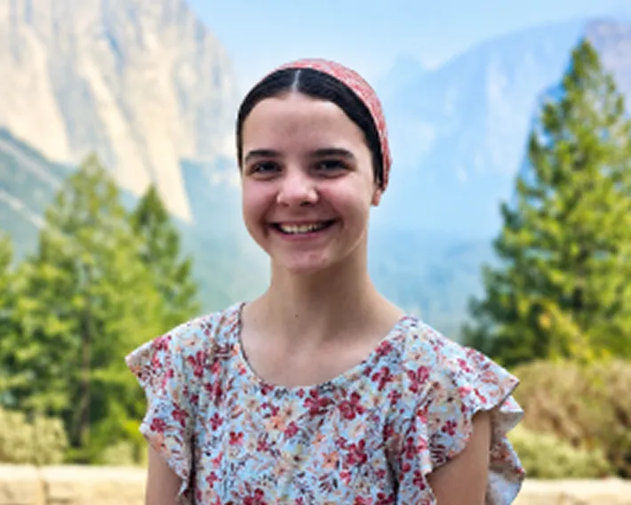 Jubilee wearing a floral blouse and a pink headband smiles outdoors with mountains and pine trees in the background.