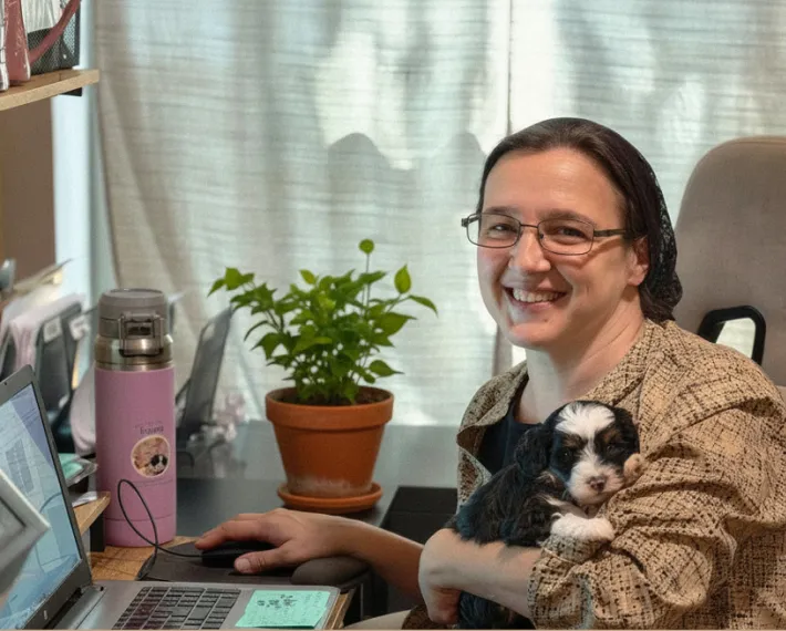 Cindy sits at a desk with a laptop, smiling at the camera while holding a small puppy.
