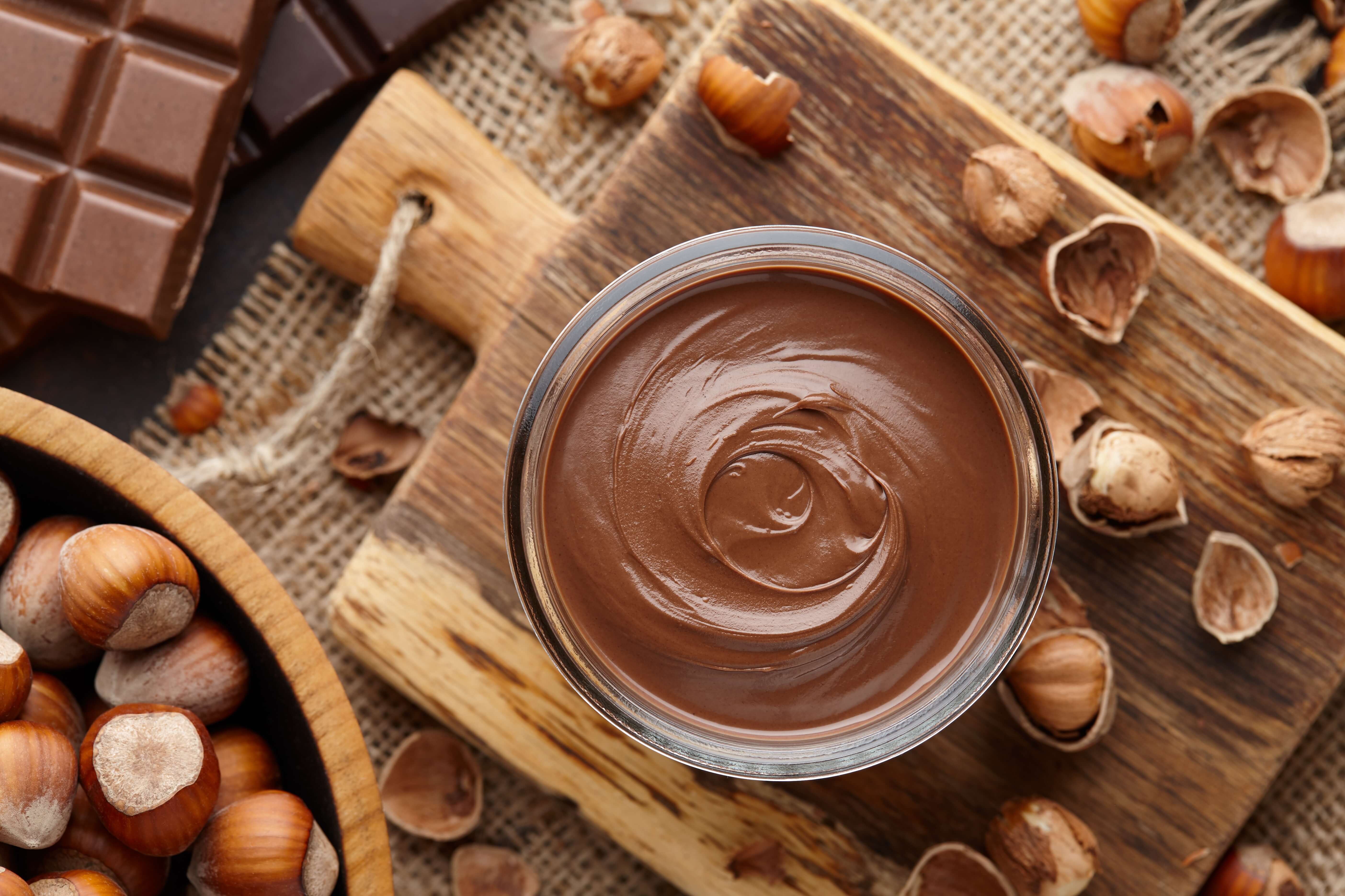 Jar of chocolate hazelnut spread surrounded by hazelnuts and chocolate bar pieces on a wooden board