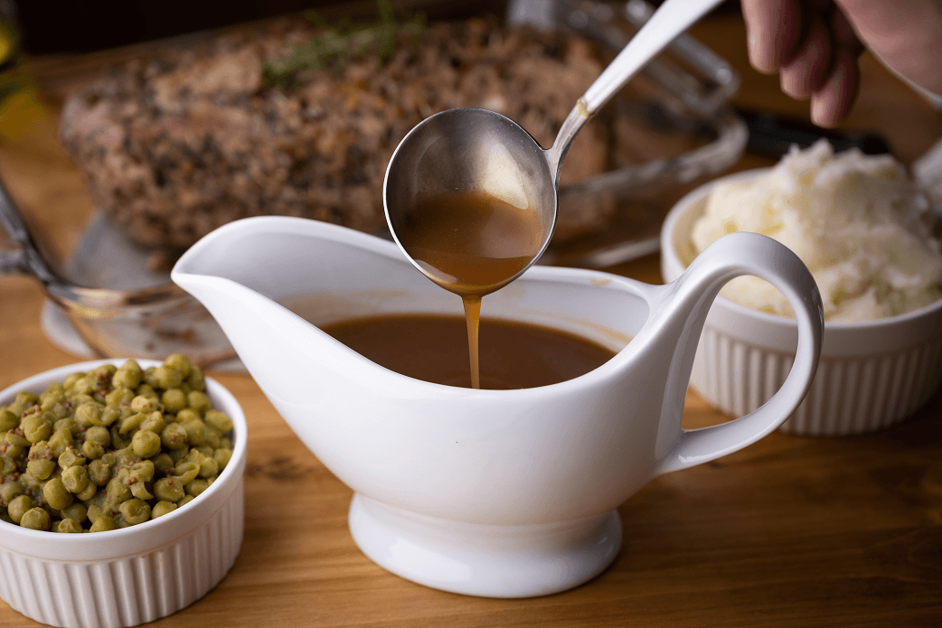 Gravy being poured from a gravy boat next to mashed potatoes and peas, with a roast in the background