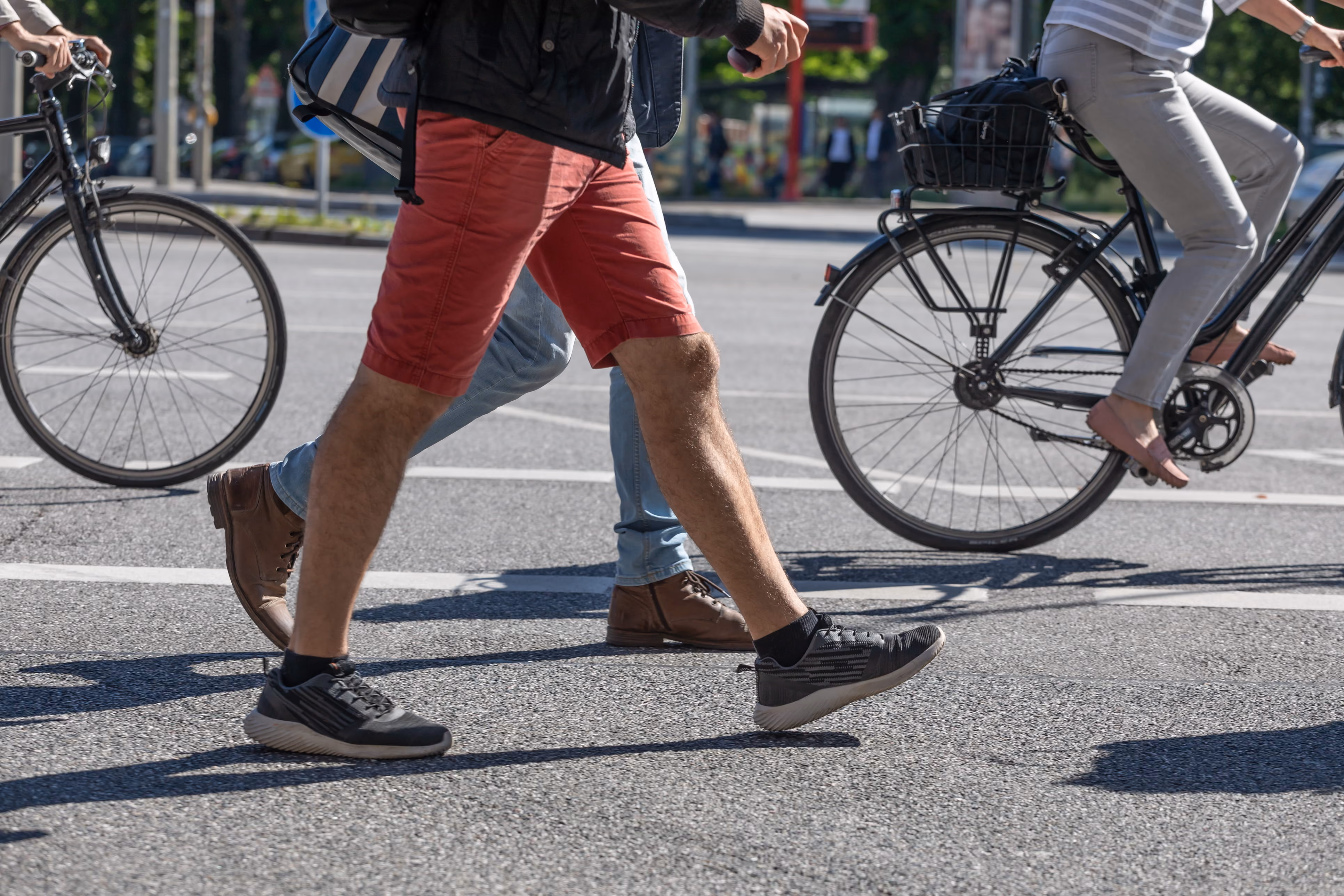 People walking and cycling across a street on a sunny day with urban greenery in background.
