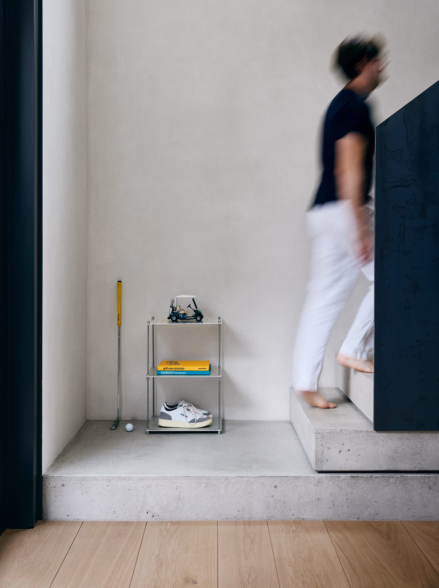 Person mit weißen Hosen und dunklem Oberteil geht barfuß eine Treppe aus Beton hinauf, links davon steht ein kleiner Metallregal mit Spielzeugauto, gelbem Buch und weißen Turnschuhen, daneben ein Golfschläger und ein Golfball vor einer weißen Wand.
