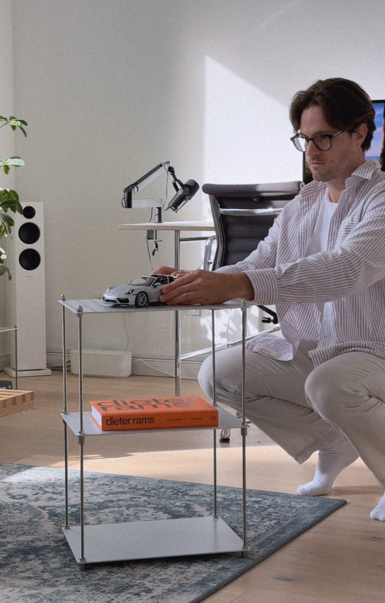 Mann mit Brille und gestreiftem Hemd kniet auf einem Teppich vor einem kleinen weißen Metallregal mit drei Ebenen, auf dessen oberster Ebene ein Modellauto steht und auf der mittleren Ebene ein orangefarbenes Buch liegt. Im Hintergrund sind ein Stuhl, ein Schreibtisch mit einer Lampe und ein Lautsprecher neben einer Pflanze zu sehen.