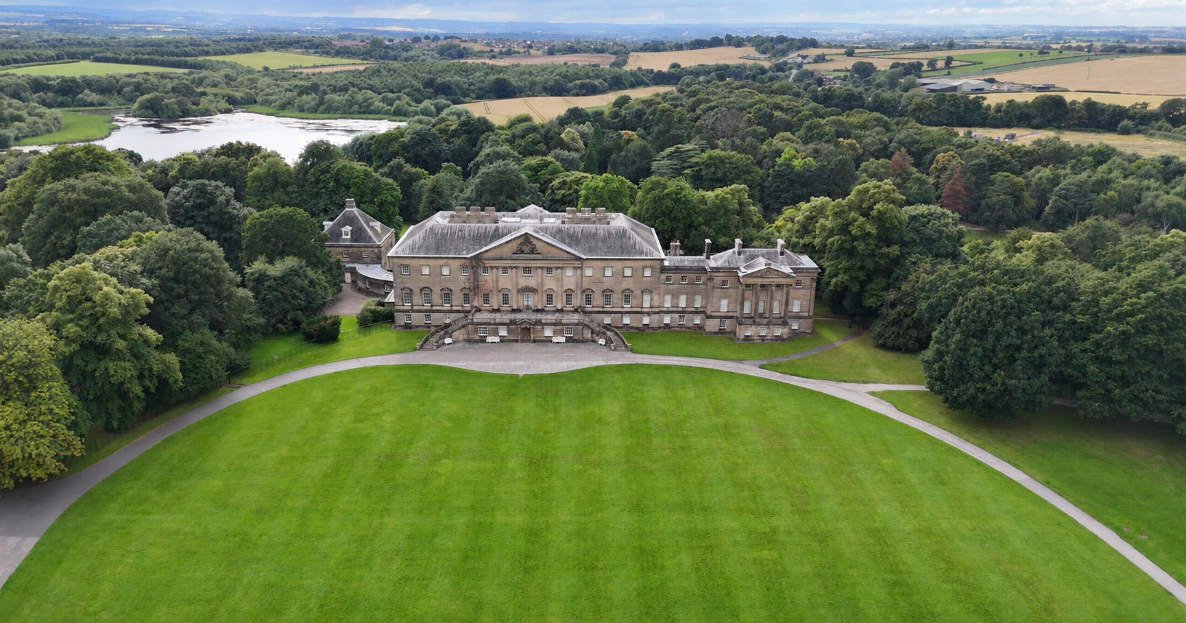 Aerial view of a grand country house surrounded by lawns, woodland and a lake.
