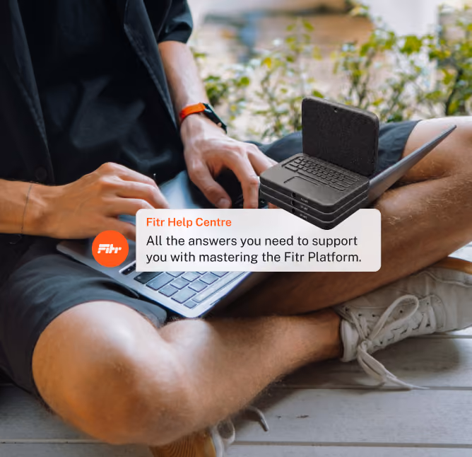 Person sitting cross-legged on the floor typing on a laptop, wearing a black shirt and shorts with a smartwatch on the wrist; overlay text box reads 'Fitr Help Centre: All the answers you need to support you with mastering the Fitr Platform.'