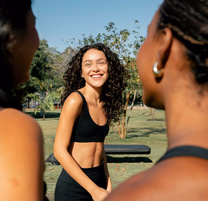 A young woman with curly hair smiling and wearing a black sports bra and leggings, standing outdoors in a park, conversing with two other people visible from behind.