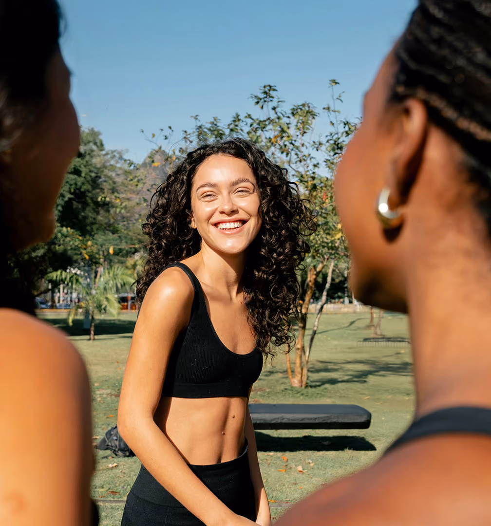 Smiling woman with curly hair in black sportswear standing outdoors in a park, facing two other women slightly blurred in the foreground.