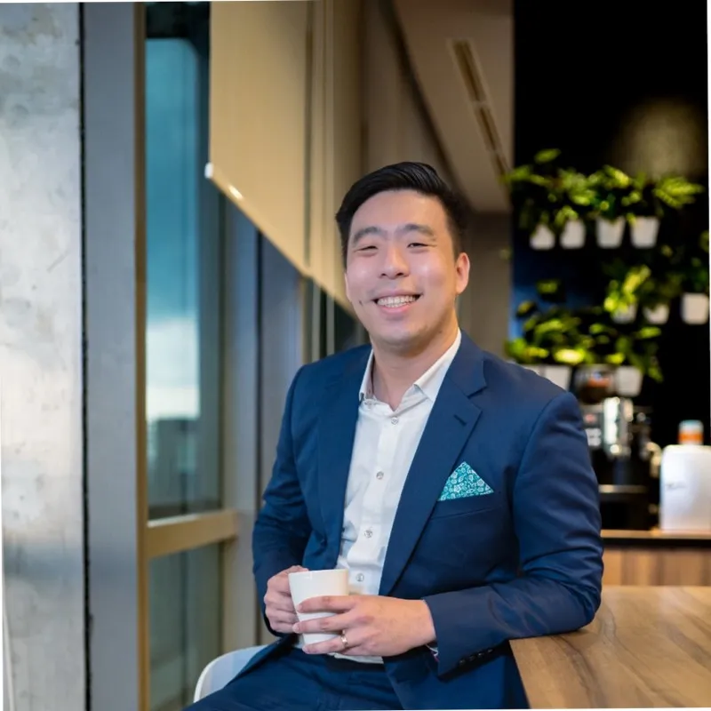 Smiling man in a blue suit and white shirt holding a cup while sitting at a wooden counter in a modern office space with plants on the wall in the background.