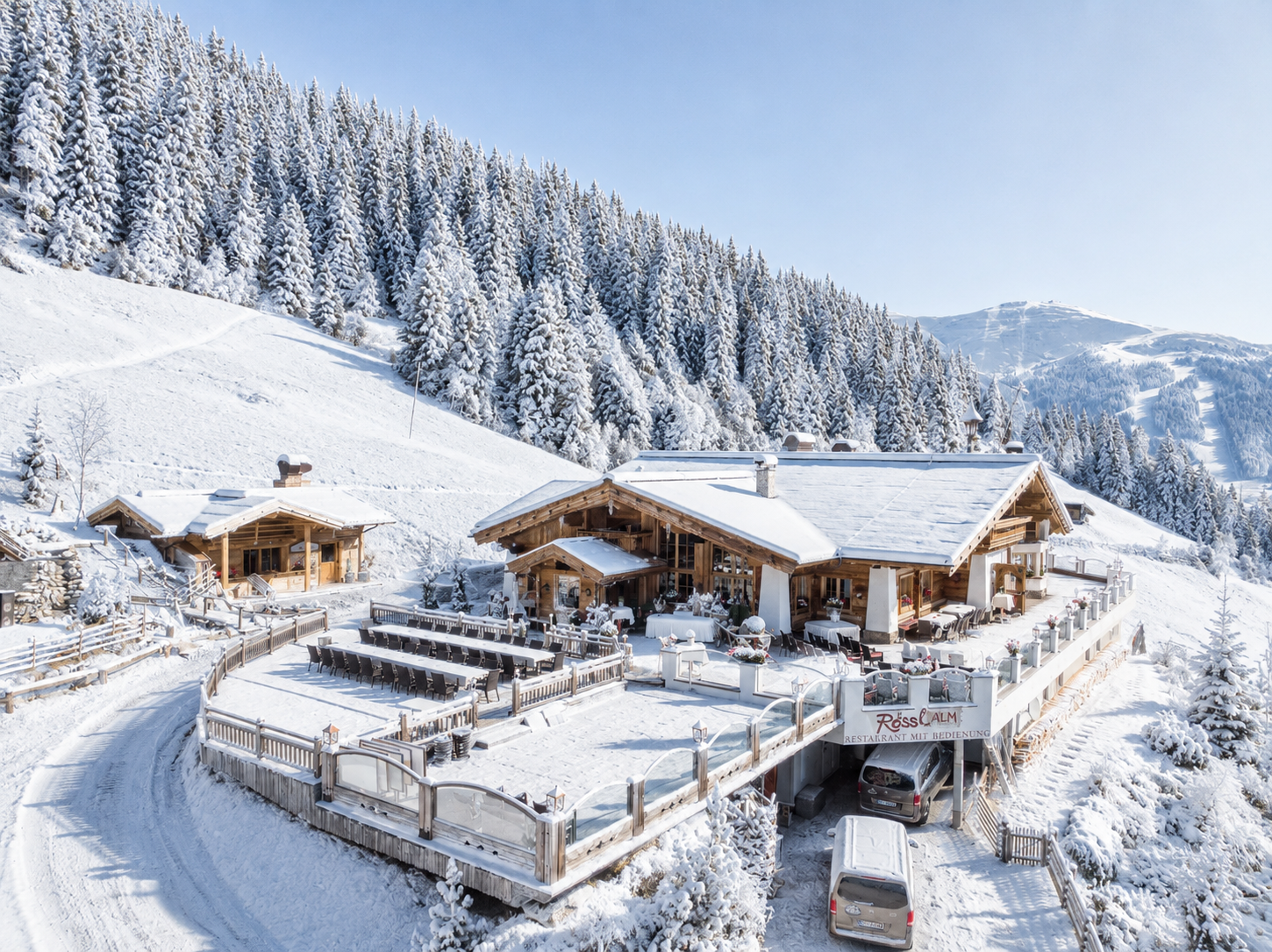 De Rössl Alm in de sneeuw: het chalet en het terras bij het middenstation, met besneeuwde dennen op de helling erachter