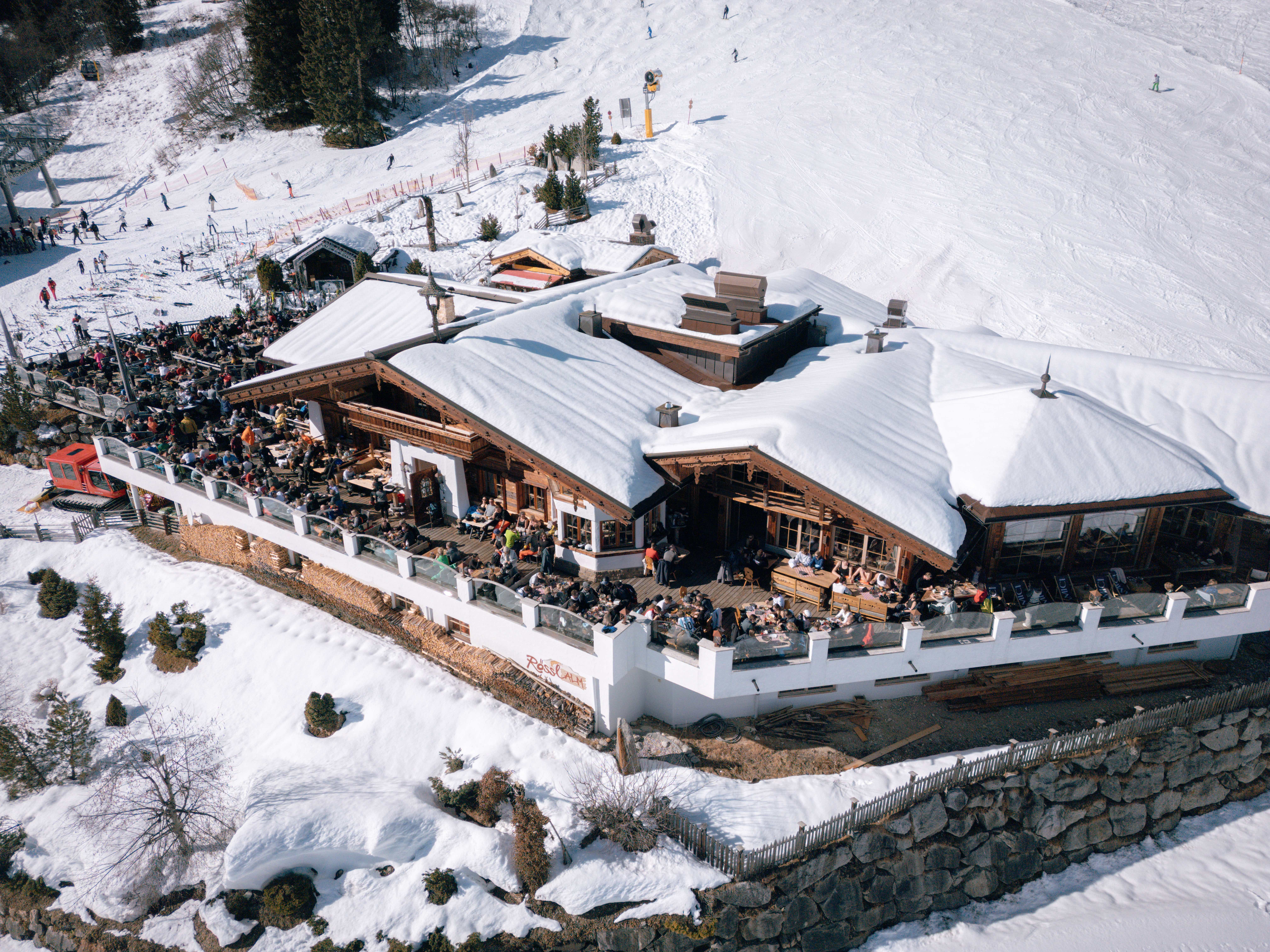 Het volle terras van de Rössl Alm van bovenaf, met de besneeuwde piste erachter