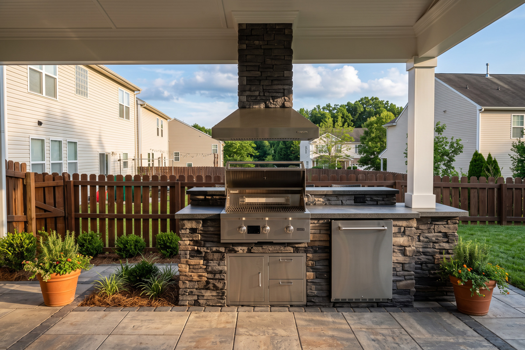 Covered outdoor kitchen with a stainless grill, stone base, and stone chimney hood on a paver patio