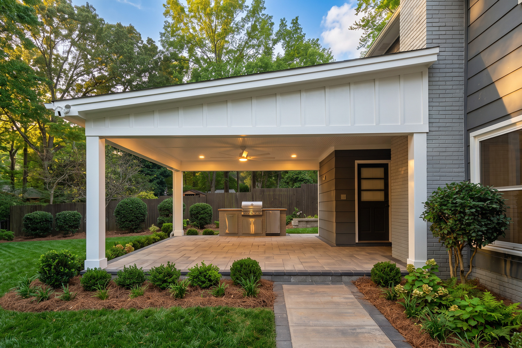 White open-sided covered patio with a peaked roof and built-in grill station, reached by a paver walkway