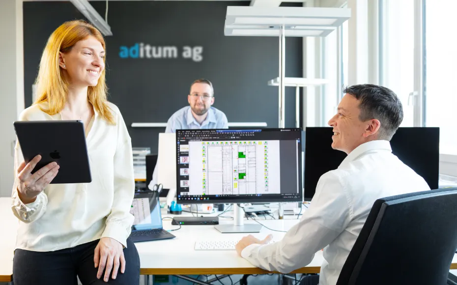 Drei Mitarbeitende in einem hellen Büro mit Computern; eine Frau hält ein Tablet und lächelt, zwei Männer sitzen und unterhalten sich, Bildschirm zeigt technische Zeichnungen, Firmenname aditum ag an der Wand.