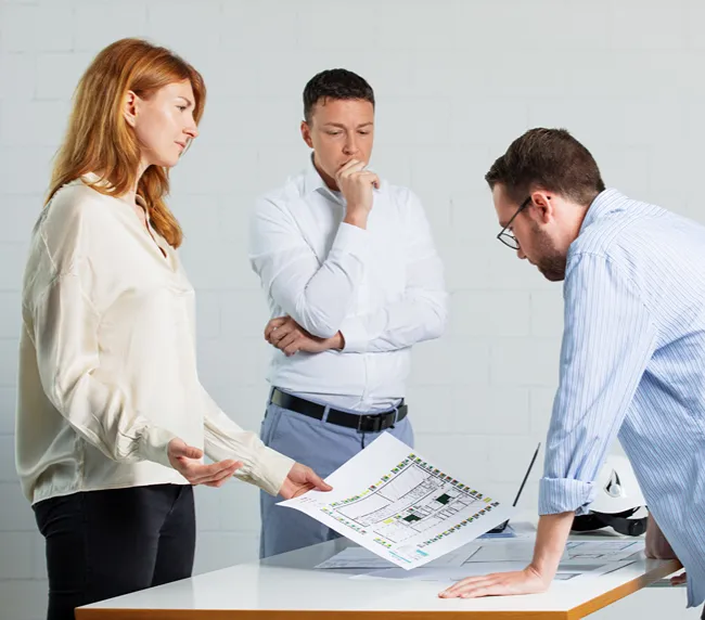 Drei Personen in einem Büro diskutieren einen Bauplan auf einem Tisch; eine Frau zeigt das Dokument, während zwei Männer aufmerksam zuhören.