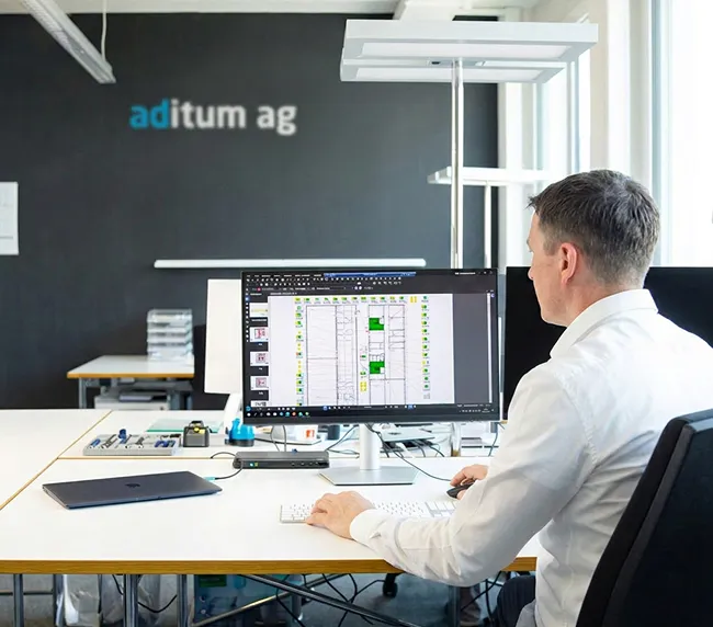 Mann in hellem Hemd sitzt an einem Schreibtisch in einem modernen Büro und betrachtet am Computerbildschirm einen digitalen Plan mit grün markierten Bereichen.