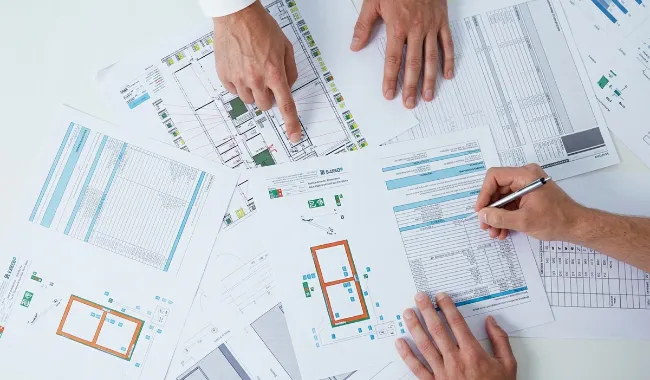 Drei Hände arbeiten gemeinsam an technischen Plänen und Tabellen, eine Hand zeigt auf ein Architekturdiagramm, eine andere hält einen Stift und schreibt auf ein Tabellenblatt.