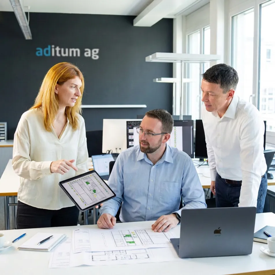 Drei Personen in einem hellen Büro mit Grossfenstern besprechen Baupläne; eine Frau zeigt einem sitzenden Mann Pläne auf einem Tablet, ein weiterer Mann steht neben ihnen und schaut zu, Laptop und weitere Unterlagen liegen auf dem Tisch, im Hintergrund Schriftzug 'aditum ag' an der Wand.