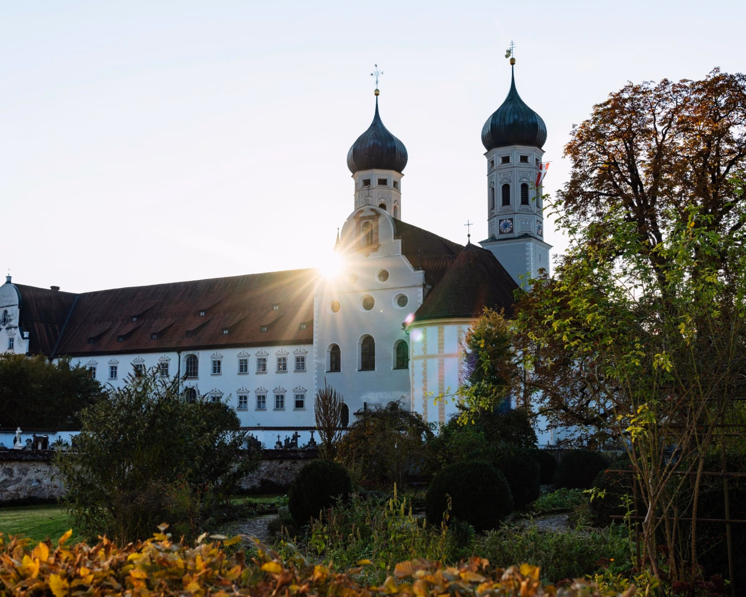 Kloster Benediktbeuern mit zwei Zwiebeltürmen im Gegenlicht, davor ein herbstlicher Garten