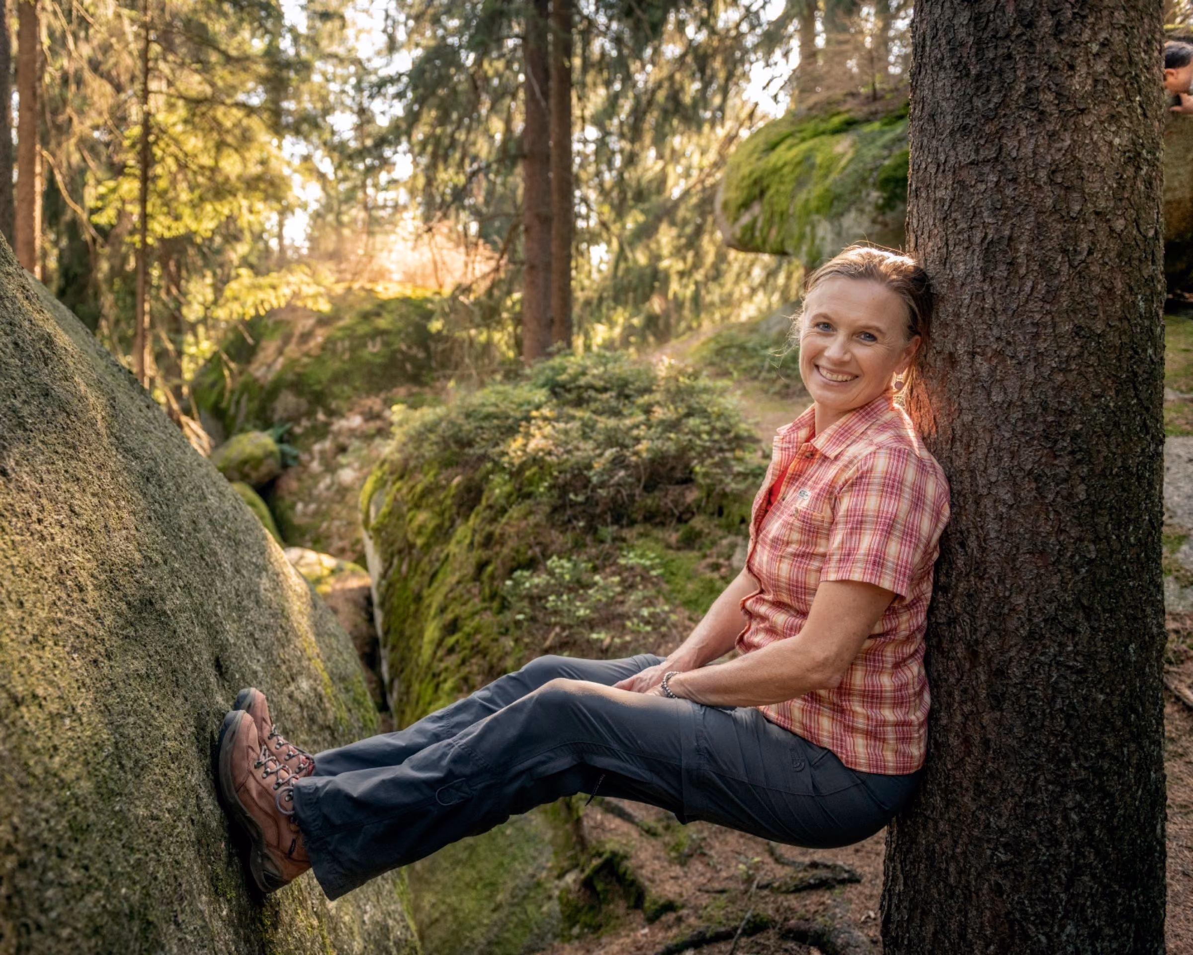 Lächelnde Frau in kariertem Hemd lehnt im Wald im Fichtelgebirge zwischen Baumstamm und moosigem Felsen
