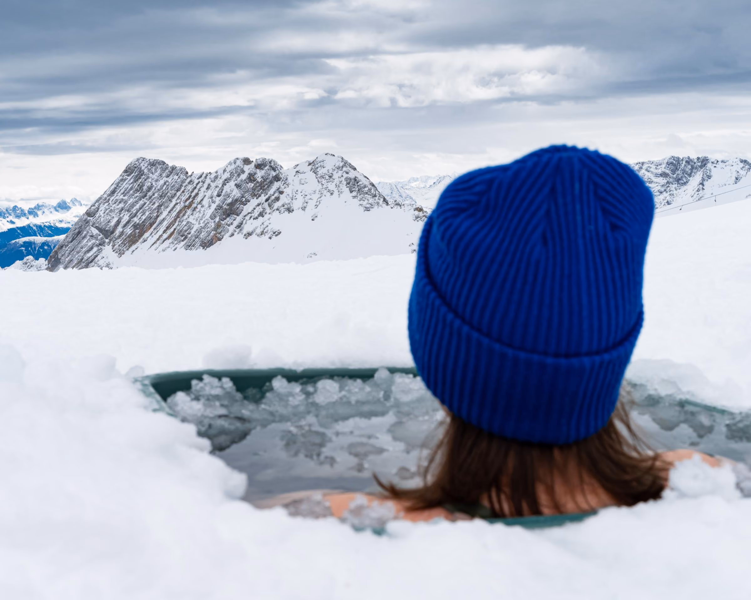 Frau mit blauer Mütze sitzt im Eisbad im Schnee auf der Zugspitze, im Hintergrund schneebedeckte Berggipfel