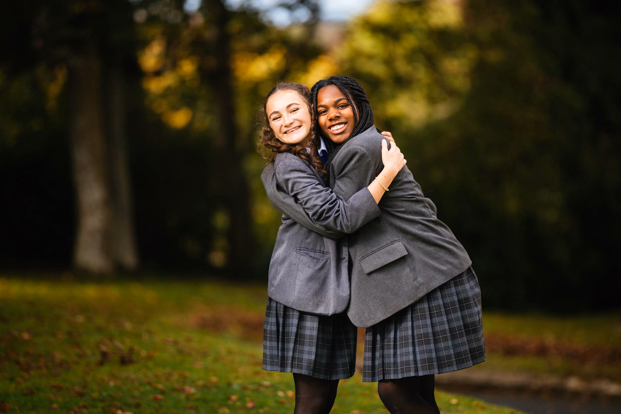 Two schoolgirls wearing grey blazers and plaid skirts hugging each other and smiling outdoors with green grass and trees in the background.