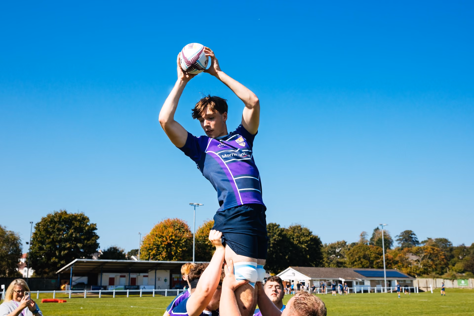 Rugby player in blue and purple uniform lifted by teammates during a lineout, catching the ball against a clear blue sky at an outdoor rugby field.