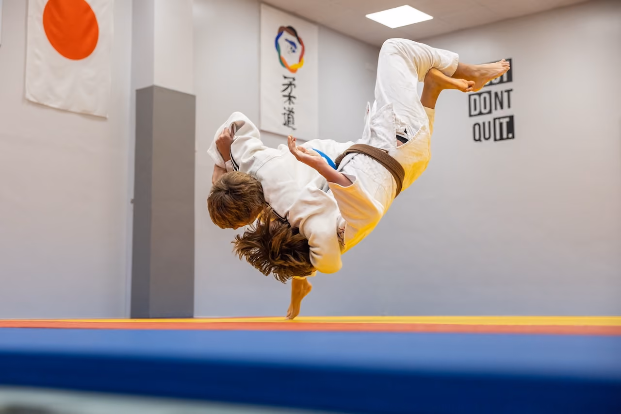 Two judokas in white gi perform a judo throw in a dojo with Japanese flag and motivational signs on the wall.