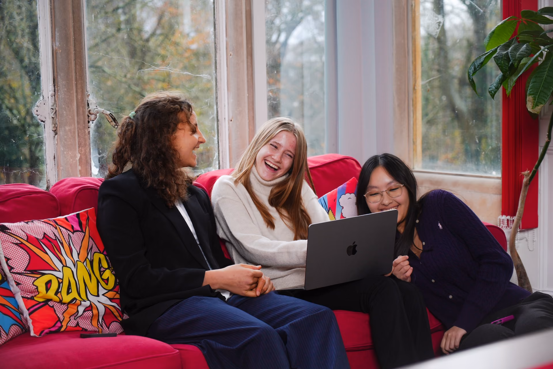 Three young women sitting on a bright pink sofa by large windows, smiling and looking at a laptop together with colourful comic-style cushions behind them.
