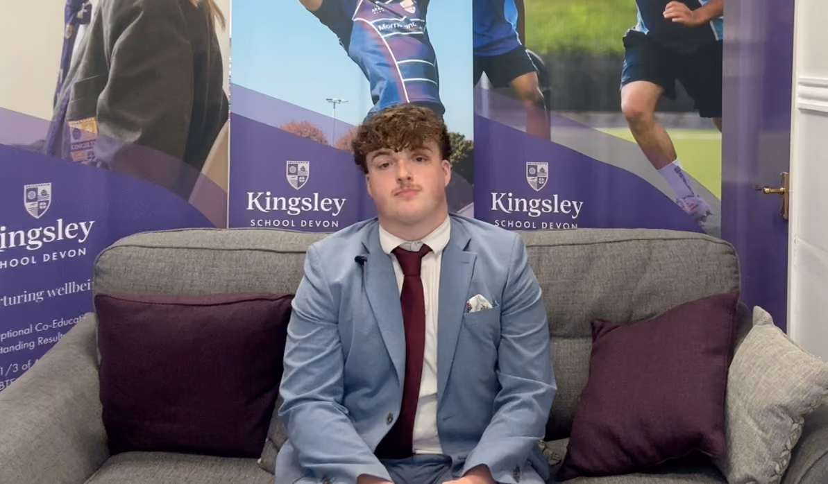 A young man with curly hair wearing a light blue suit, white shirt, and maroon tie sits on a grey couch with purple cushions in front of Kingsley School Devon banners featuring students playing sports.
