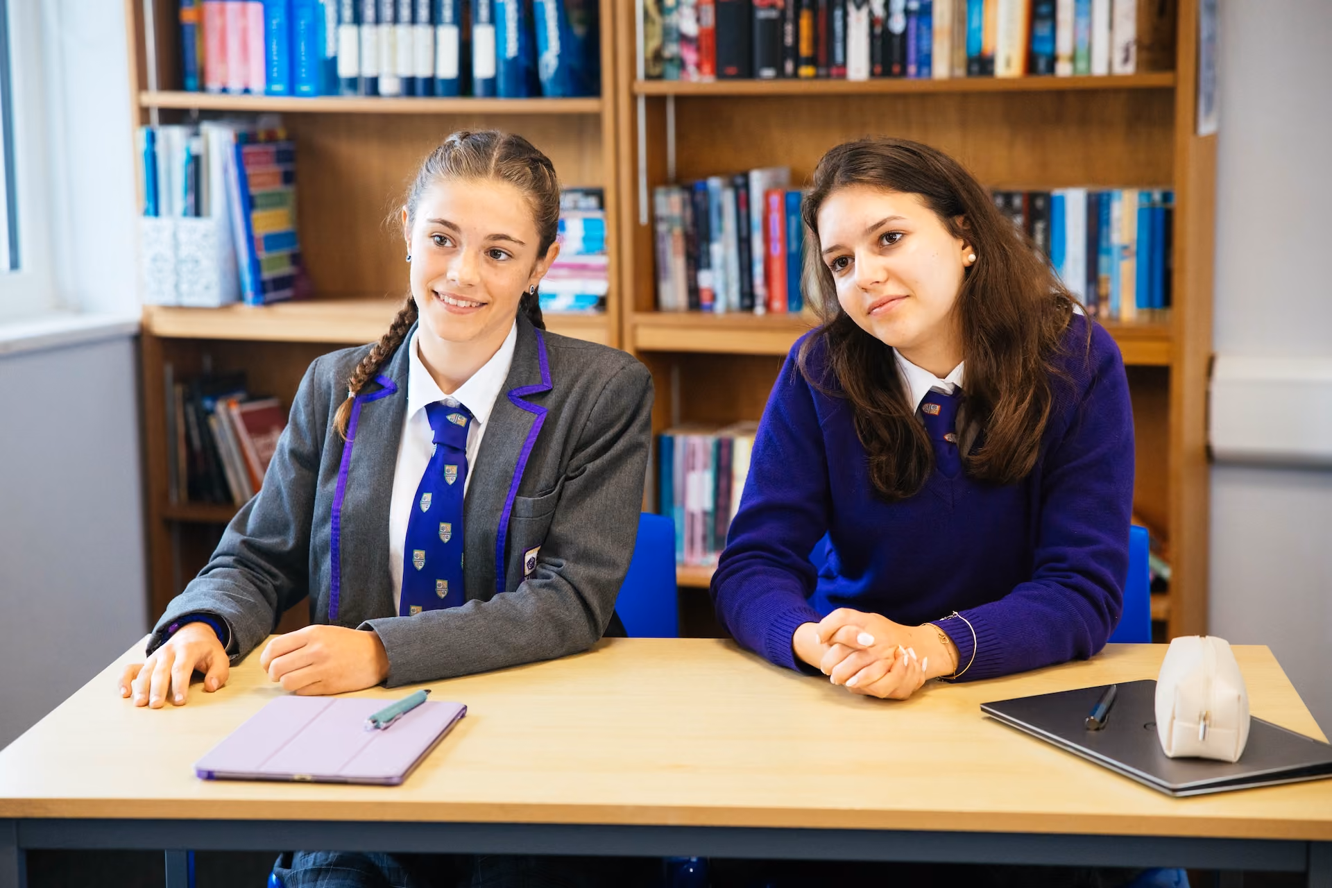 Two teenage girls wearing school uniforms sitting at a desk in a library with bookshelves in the background, both looking slightly to the side and smiling gently.