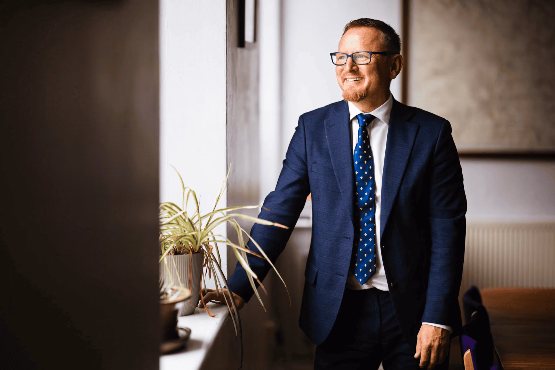 Man with glasses wearing a navy blue suit and patterned tie stands by a window smiling, with a potted plant on the windowsill.
