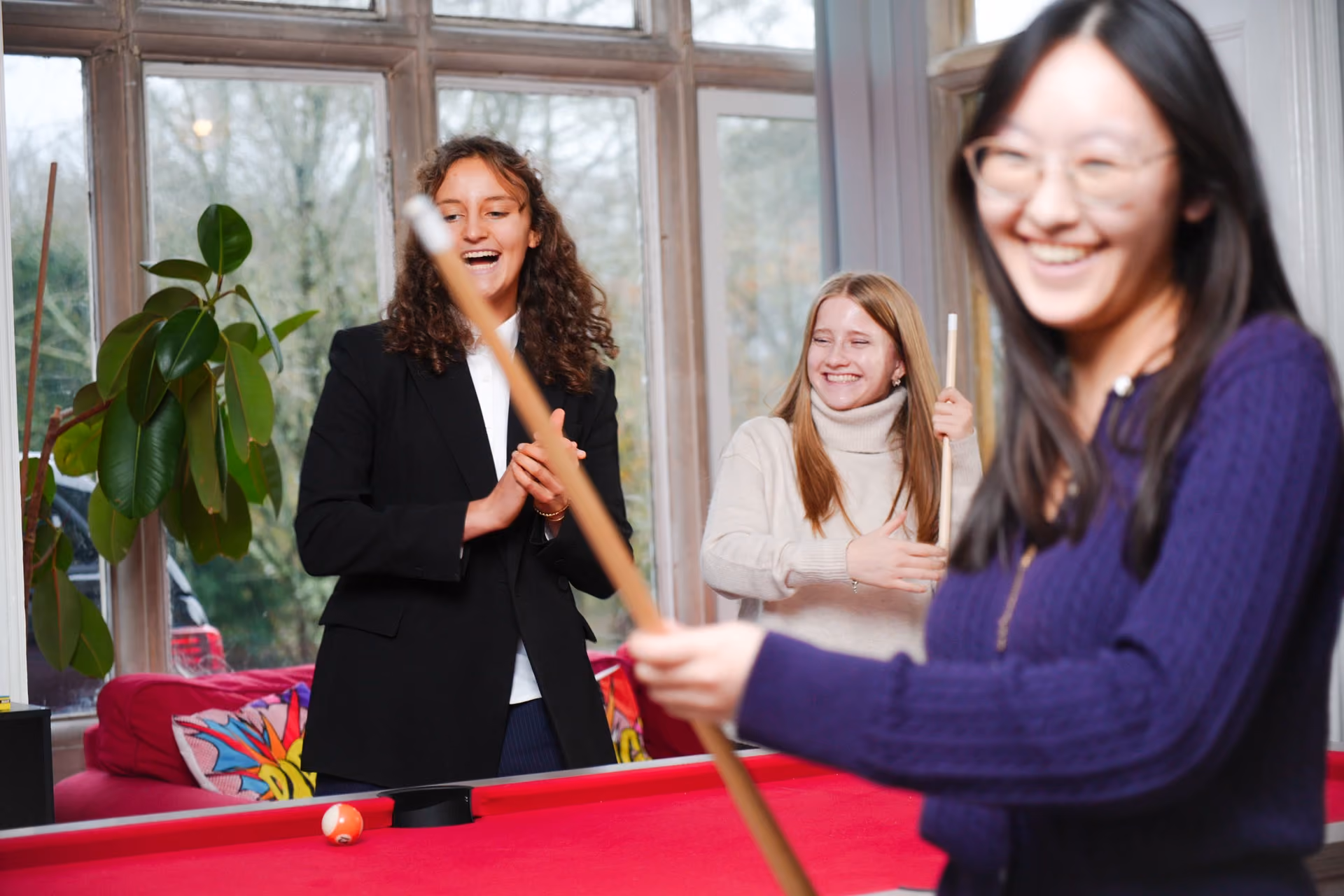 Three young women smiling and enjoying a game of pool with a bright red pool table indoors near large windows and a houseplant.