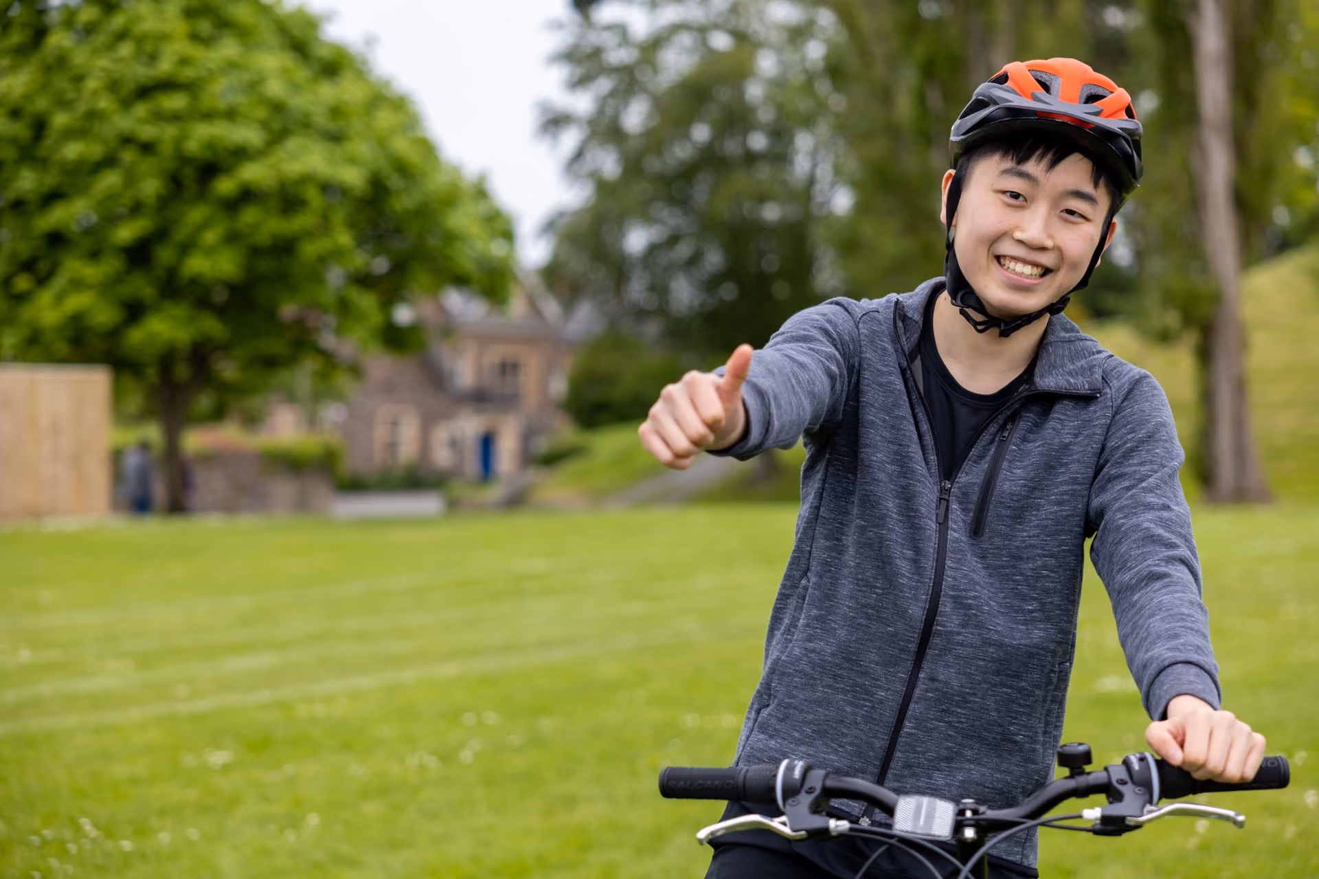 Smiling young person wearing a red and black helmet and grey zip-up jacket, sitting on a bicycle in a green park showing a thumbs-up gesture with their right hand.