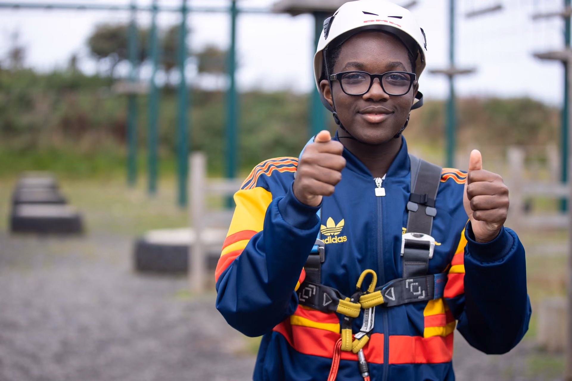 Teenager wearing a helmet, glasses, and a blue Adidas jacket with red and yellow stripes giving a double thumbs-up while secured in climbing harness outdoors.