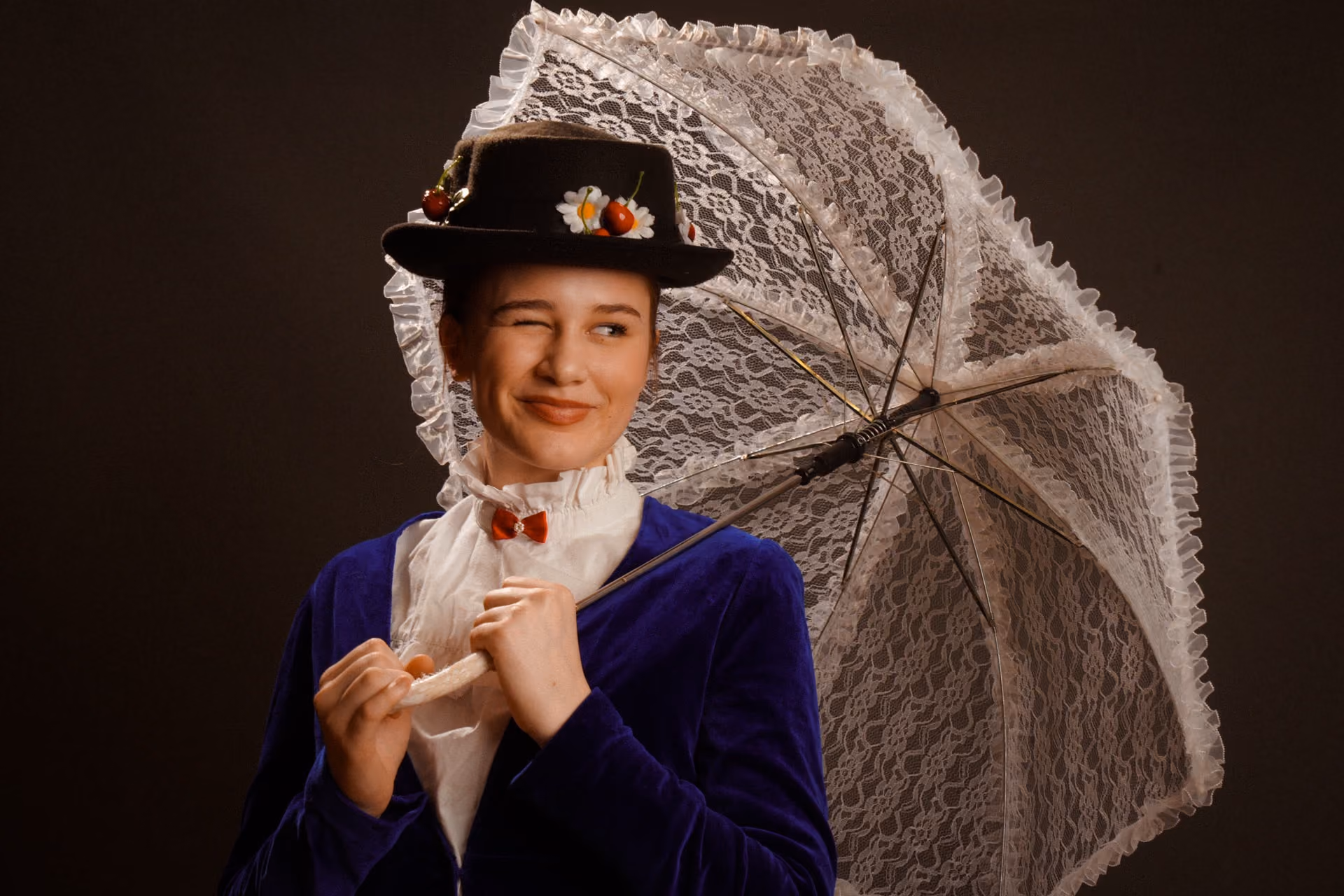 A woman dressed as Mary Poppins winks while holding a white lace parasol and wearing a black hat decorated with flowers and cherries, a white blouse with a red bow tie, and a dark blue jacket.