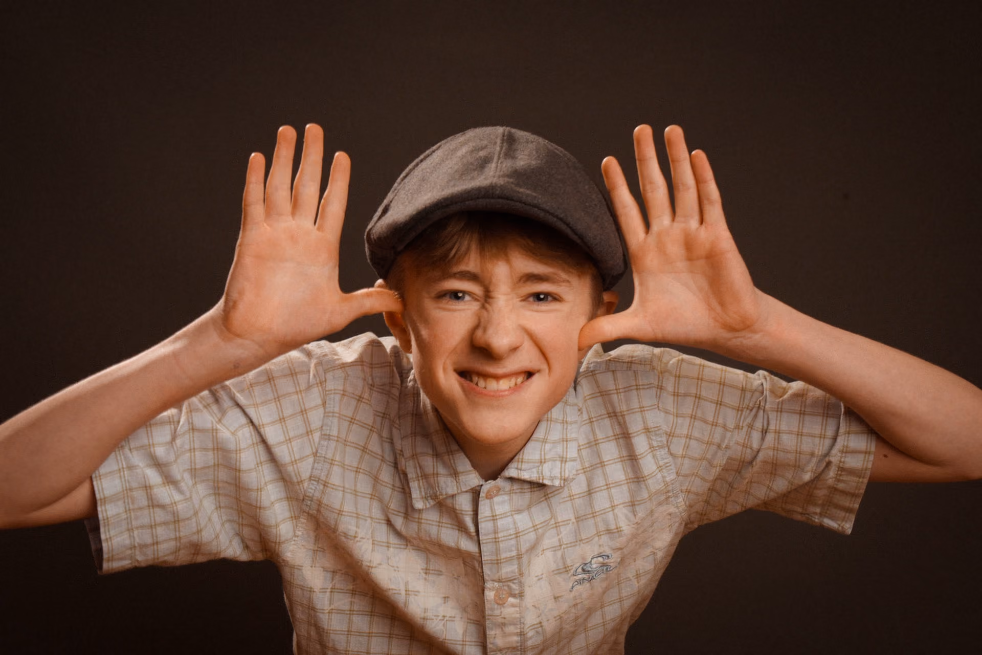 Young boy wearing a flat cap and a checked short-sleeve shirt making a playful face with both hands raised beside his head, fingers spread wide, and smiling at the camera against a dark background.