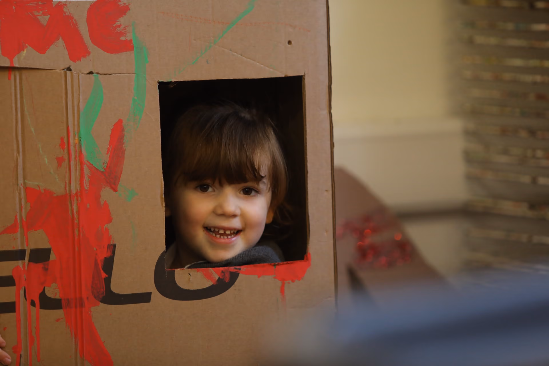 Smiling young child with brown hair peeking through a square cutout in a large cardboard box decorated with red and green paint.
