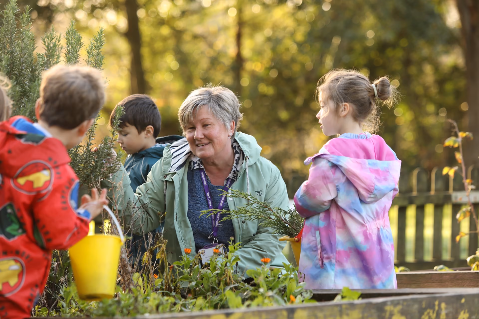 Woman in a green jacket gardening outdoors with three children, two boys and one girl, surrounded by plants and flowers during daylight.
