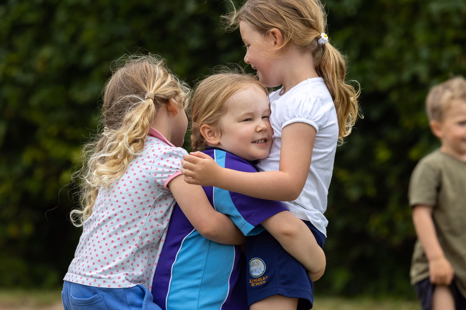 Three young children outdoors hugging and smiling, with green foliage in the background and a boy blurred in the right background.