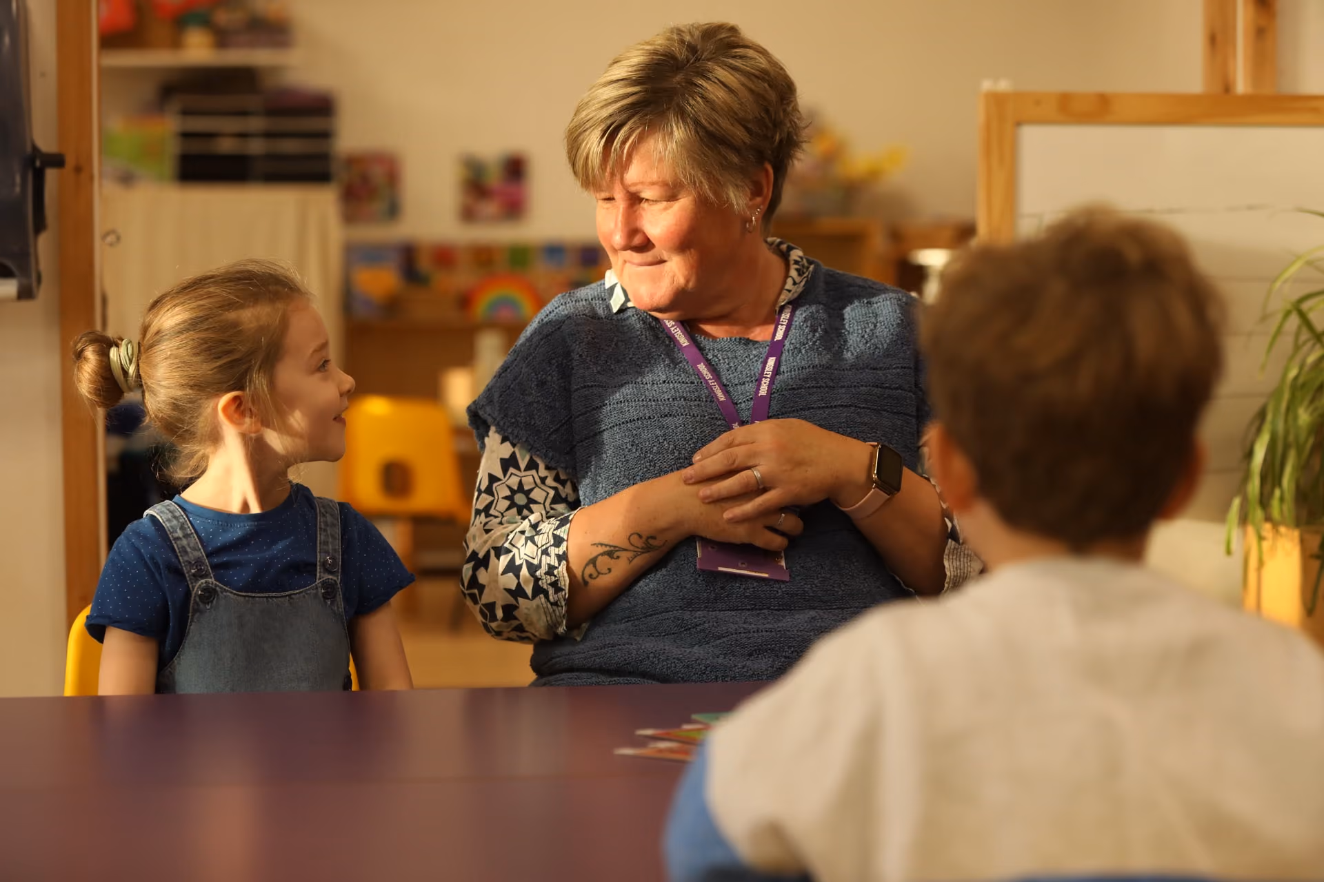 A woman with blonde hair and tattoos on her arm smiles and interacts warmly with a young girl wearing a blue shirt and denim overalls, while a young boy with short hair sits in the foreground in a classroom setting.
