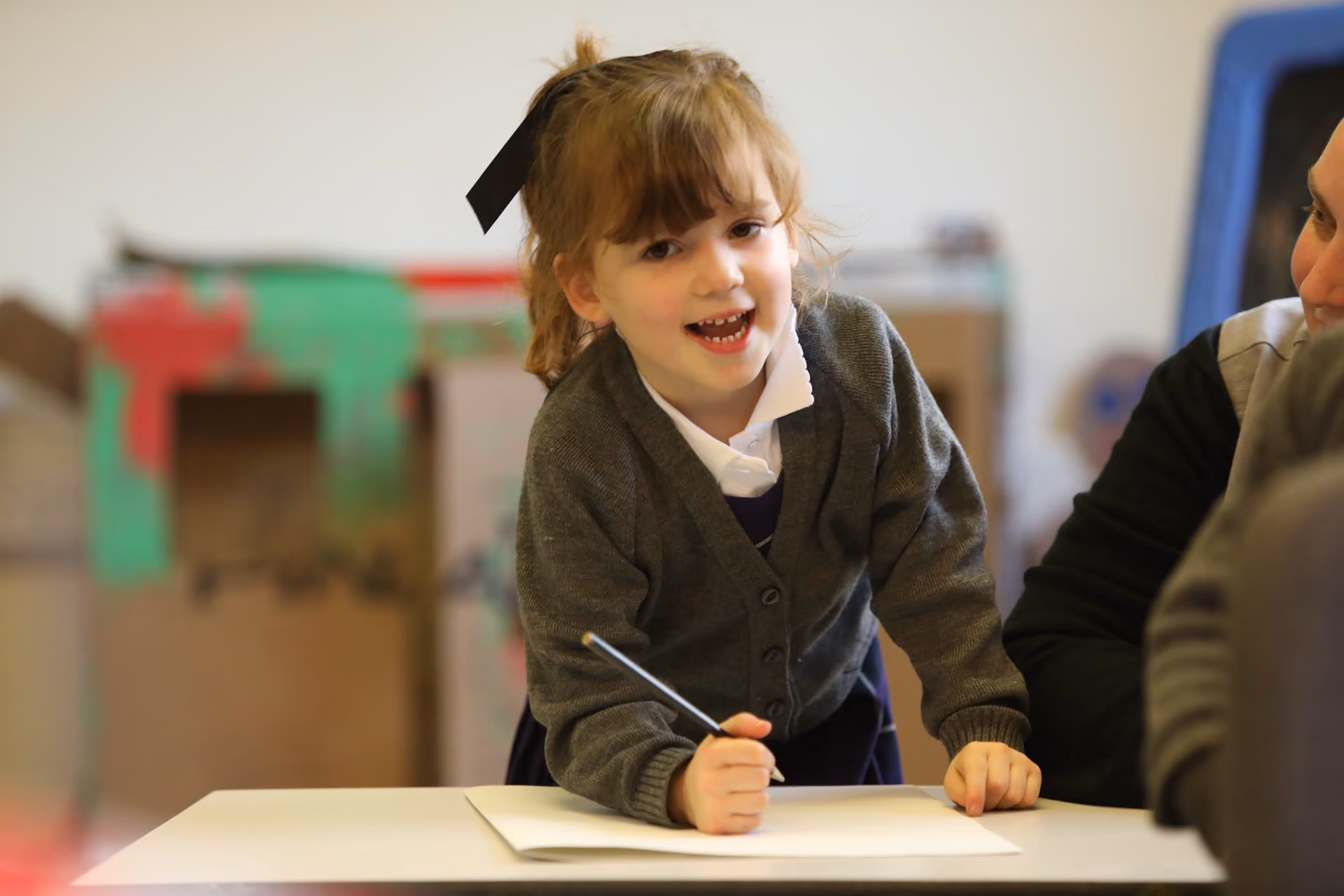 Young girl with a black bow in her hair, wearing a grey cardigan and white collared shirt, smiling and writing with a pencil on a sheet of paper at a table.